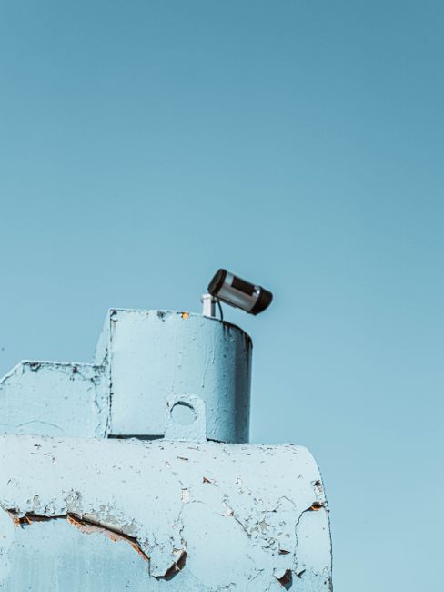 Vertical shot of a surveillance camera on a rusty, light blue industrial rooftop with a clear sky.